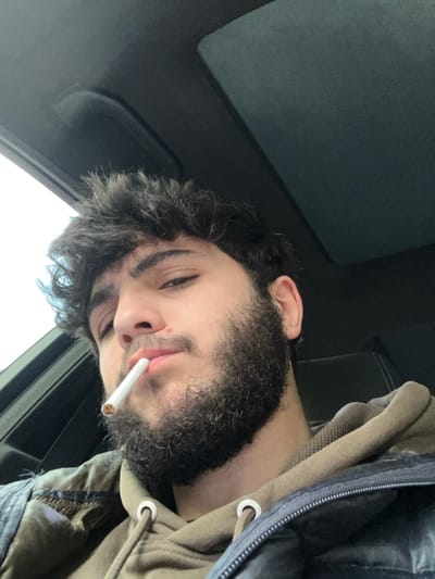 A close-up, slightly low-angle photograph taken inside a vehicle in As-Salihiya Municipality, Syria, depicts a young adult male. The subject has dark, curly hair and a full, dark beard. A small reddish mark or mole is visible on his right upper lip. He wears an olive green hooded sweatshirt underneath a dark, likely black or navy blue, zippered puffy jacket. A white cigarette with a light brown filter is held between his lips, pointed slightly downwards; it appears unlit or inactive. The background reveals the dark fabric headliner of a car's roof, with a portion of a sunroof or overhead console structure visible. A faint light source, consistent with an overcast sky, is visible through a window or sunroof opening in the upper left of the frame. A dark headrest of a car seat is partially discernible to the right of the subject's head. The subject looks directly towards the camera with a neutral expression.