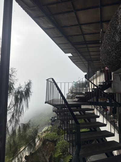 The image depicts an outdoor seating area with a view of a misty mountainous landscape, likely in Kintamani, Indonesia. A prominent feature is a dark metal spiral staircase with wooden steps, leading upwards and to the right. To the left, a large, dark vertical beam frames the scene, and a tree with wispy, drooping branches extends into the frame. Beyond the railing of the staircase, a lush green hillside is visible, shrouded in thick fog, with some wooden pathways and scattered vegetation. The upper portion of the image shows the underside of a roof structure, supported by dark metal beams, with a large woven decorative object hanging from the ceiling on the right. A few indistinct figures can be seen in the background near the stairs.
