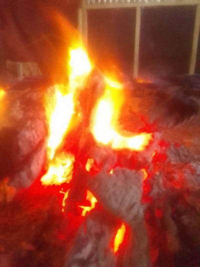 A blurry, close-up shot displays a vibrant bonfire. Intense orange and yellow flames rise from the pile of wood, illuminating surrounding gray ash and embers. A faint structure with windows is visible in the background, suggesting an outdoor setting at night. The fire's light casts a warm glow, creating an atmosphere of heat and energy.