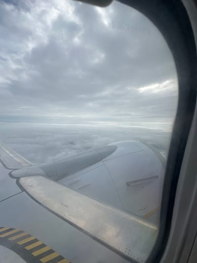 An aerial view from an airplane window shows a vast expanse of clouds below and an overcast sky. The wing of the aircraft, made of metal panels with visible rivets, extends into the frame, featuring a black and yellow striped anti-slip marking. Part of an aircraft engine nacelle is also visible. The scene is dominated by shades of gray and white, with diffused sunlight breaking through the clouds. No people are visible. There is no indication of Krivina, Bulgaria.