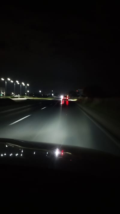 The image depicts a nighttime view from inside a vehicle traveling on a highway, possibly near Graf Ignatievo, Bulgaria. The road is wet, reflecting the headlights of the vehicle and the taillights of a car ahead. Streetlights are visible along the left side of the road, illuminating the roadside. The scene is dark, with the primary light sources being the vehicle's headlights and the distant lights of other vehicles and infrastructure. The view is somewhat blurred, suggesting movement.