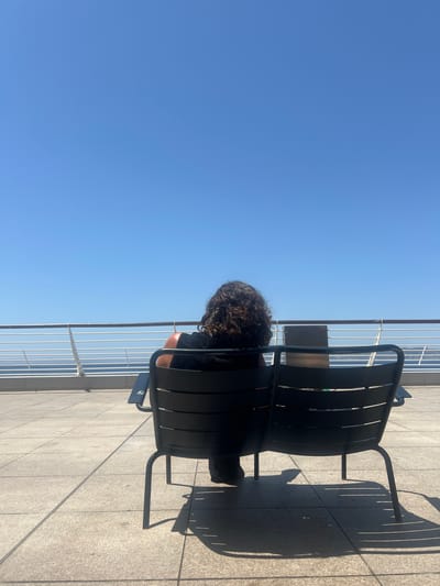 A person with curly brown hair is sitting in a black chair on a tiled patio, facing away from the camera. They are wearing a black top. The person is seated at a table. In the background, a railing separates the patio from a body of water and the sky. The sky is clear and blue. The scene appears to be outdoors on a sunny day. The location is a city terrace in Marseille, France, likely overlooking the Mediterranean Sea.