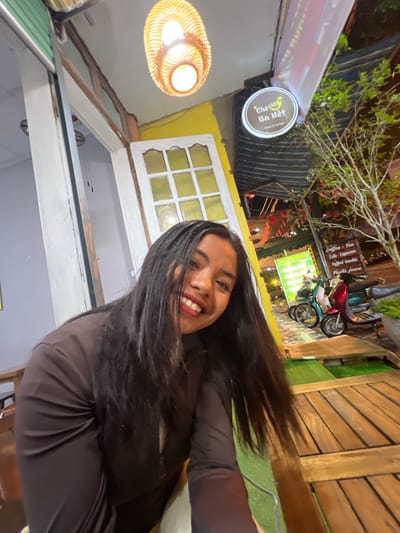 A young woman with long, dark hair and a brown long-sleeved shirt smiles at the camera. She is seated at a wooden table outdoors, with a thatched light fixture hanging above her. Behind her, there is a yellow building with a white-paned window, a sign that reads "Chè Bà Nét - Food & Drink," and a rack of menus advertising coffee, tea, smoothies, and fruits. Several motorbikes are parked to the right of the scene, and a tree with red leaves grows in the background. The overall impression is that of a casual outdoor dining setting in Bắc Hà, Vietnam.