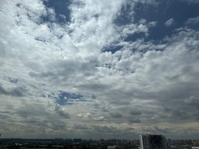 The image shows a cloudy sky over the city of Lyubertsy, Russia. The sky is filled with a mix of white and grey clouds, with some patches of blue visible. The clouds are varied in thickness and texture, with some appearing wispy and others more dense and cumulus-like. The horizon shows a cityscape with numerous buildings of varying heights, including residential blocks and possibly some commercial structures. Trees are visible in the foreground, indicating a suburban or park-like area within the city. The overall impression is that of a slightly overcast day.