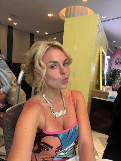A woman with blonde hair and a chain with letters "PRIME" is looking at the camera with pursed lips, possibly taking a selfie. She is wearing a tank top with a colorful graphic design and pink straps. The background appears to be a public space, possibly a restaurant or store, with yellow fixtures and recessed lighting. An "amazon" box and plants are seen in the background. The location is likely within the city of Miami Beach, Florida, based on the presence of "amazon" and "Prime" and style of the establishment. Other people, including those wearing casual clothing, are visible in the background.