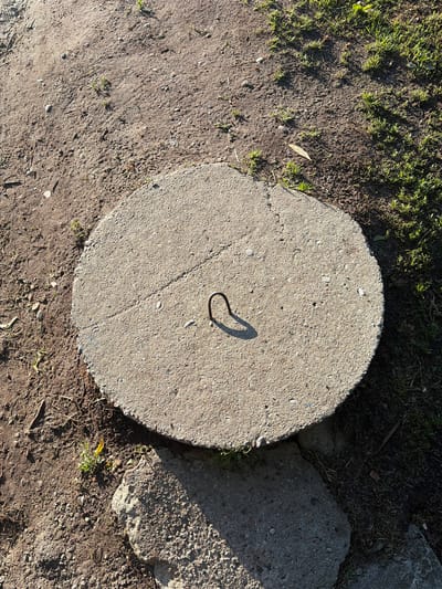 The image depicts a circular concrete manhole cover partially submerged in soil and grass. The manhole cover has a metal loop protruding from its surface, casting a shadow. The surrounding environment consists of dirt paths, patchy grass, and some scattered pebbles. The lighting suggests it is daytime with the sun casting distinct shadows. There are no people or animals visible in the image. The scene appears to be an outdoor, urban or suburban setting, likely a park or a less maintained area within a city. Based on the provided context, the image is located in Kaliningrad, Russia.
