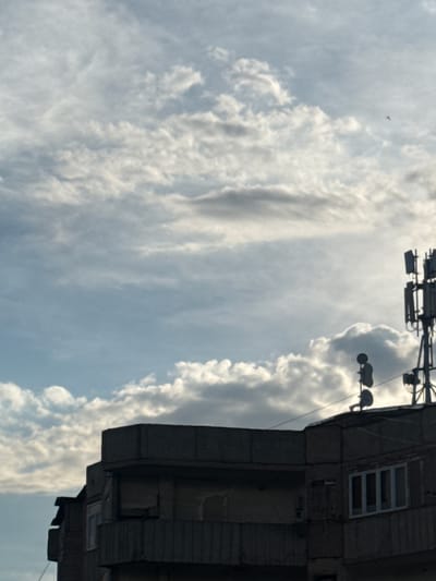 The image shows the upper portion of a concrete building against a cloudy sky. The building has balconies with railing and multiple windows. On the roof of the building, there are communication antennas and dishes. In the distance, a small object, possibly a bird or a drone, is visible in the sky. The lighting suggests it is either early morning or late afternoon. The scene is consistent with an urban environment, potentially in Abovyan, Armenia, given the architectural style and the presence of communication infrastructure commonly found in cities.