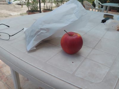The image displays a close-up of an outdoor table setting, likely a patio or garden in Silistra, Bulgaria, during bright daylight. The main subject is a light-colored, possibly beige or off-white, plastic table with a tiled surface pattern.
On the table, several items are arranged. A vibrant red apple with a short brown stem sits prominently towards the right side of the frame. To its left, a crumpled white paper bag, possibly containing a snack or sandwich, takes up a significant portion of the table space. Further to the left, a pair of black-framed eyeglasses rests near the table's edge. Towards the upper right, a dark wallet or small pouch is visible, alongside a circular dark object that could be a coaster or a small tin, and some clear plastic packaging.
The background shows a relaxed outdoor environment. Potted plants with green foliage are visible, creating a natural backdrop. White plastic chairs and another white table can be discerned further back, suggesting a larger outdoor dining or seating area. The ground beneath the table appears to be tiled. The bright, clear lighting indicates it is daytime. There are no people visible, and no discernible text on any of the objects. The scene conveys a sense of a casual, quiet break outdoors.