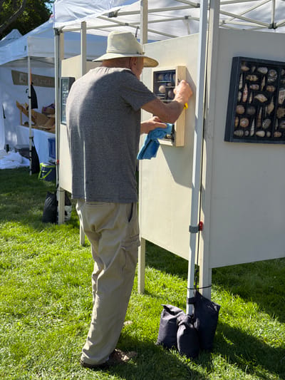 An elderly Caucasian male wearing a straw hat, a striped grey t-shirt, and khaki cargo pants is cleaning a display board at an outdoor market. He is holding a yellow scraper in his right hand and a blue cloth in his left hand, and appears to be wiping the display board. The man is standing on a grassy area with a white tent structure and another display board featuring seashells to his right. The display board he is cleaning has small compartments containing shells. In the background, other tents and display items are visible. The scene is set outdoors, illuminated by sunlight. This appears to be an art or craft fair in the Village of Sag Harbor, USA.