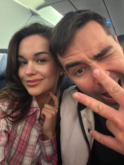 The image shows a selfie taken by a man and a woman inside an airplane. The man is positioned above the woman, and both are looking at the camera. The man is making a "peace" sign with his fingers, winking, and sticking out his tongue. He is wearing a white jacket over a dark shirt. The woman is smiling and wearing a pink and white plaid shirt. They are seated on an airplane. The location is likely in the vicinity of Емельяново (аэропорт), Russia.