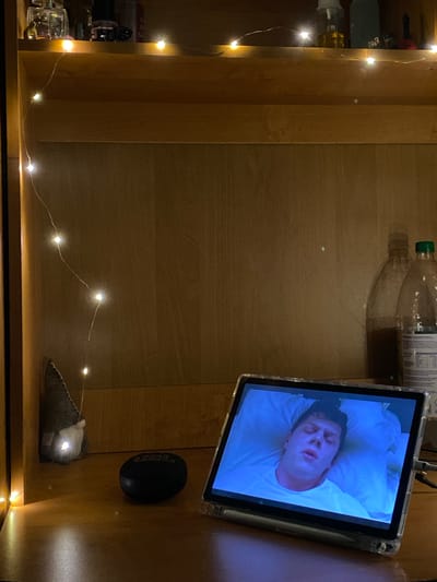 The image depicts a wooden shelf in what appears to be a dormitory or bedroom setting. A string of fairy lights with warm yellow bulbs is draped across the shelf, casting a soft glow. On the top shelf, several items are visible, including perfume bottles, hair clips, and other small objects. Below this, on the main shelf, a tablet computer is displaying a close-up of a person's face, appearing to be asleep or unconscious, with a blue-tinted lighting effect. To the left of the tablet, a small, gray gnome-like figurine with a pointed hat and white beard is illuminated by a fairy light bulb. In front of the gnome, a black, oval-shaped speaker with the "JBL" logo is present. To the right of the tablet, two large clear plastic bottles are visible, one containing a dark liquid and the other filled with a lighter liquid, with a green cap on the latter. The overall environment suggests a cozy, personal space, possibly late at night or in a dimly lit room, with the fairy lights adding to the ambiance. The location is not specified as Ivano-Frankivsk, Ukraine, and there are no indications within the image to support this claim.