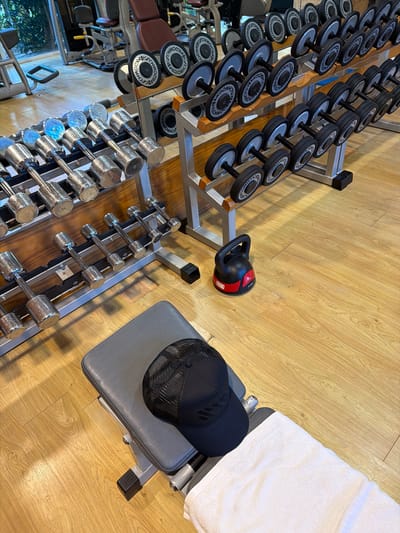 The image shows a gym setting with various exercise equipment. In the foreground, a row of chrome dumbbells is neatly arranged on a rack. To the right of the dumbbells, a gray weight bench is visible, partially covered with a white towel. A black baseball cap with a mesh back is placed on the bench. In the middle ground, a black kettlebell with red accents rests on the wooden floor. Behind the kettlebell and to the left, multiple racks of dumbbells are organized, with some displaying the brand name "TECHNOGYM". The floor is made of light-colored wood planks. The overall scene suggests a clean and organized workout space, likely a gym. The lighting indicates it could be daytime, with shadows cast by the equipment. There are no people visible in the image. The location context provided is La Gaspara, Spain.