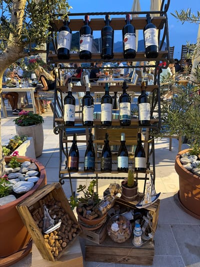 This image depicts an outdoor seating area at a restaurant in Petrovac centar, Montenegro, likely during the evening. The foreground features a metal wine rack displaying numerous bottles of wine, primarily red, on multiple shelves. The wine bottles have various labels, some with Cyrillic script and others in Latin. Below the wine rack, several wooden crates are arranged. One crate is filled with wine corks and contains a decorative miniature sailboat. Other decorative items, including a lighthouse figurine and glass spheres filled with corks and shells, are also visible. Potted plants, including an olive tree and a succulent, are placed around the seating area, and large terracotta planters with decorative stones and flowers are present. In the background, diners are seated at tables, some of which are lit by ambient lighting. The atmosphere suggests a relaxed dining experience, with the twilight sky visible.
