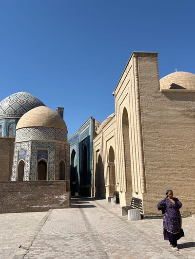 In Samarkand City, Uzbekistan, a woman stands in an open courtyard paved with light-colored hexagonal and rectangular stones. She is wearing a dark purple, long-sleeved dress with gold and blue floral patterns and a matching headscarf. Her stance is casual, with one hand on her hip and the other near her chest.
Behind her, a towering, light-brown brick building with arched entrances runs along the right side of the frame, casting long shadows. To the left, several mausoleums with ornate blue and turquoise tiled domes rise against a clear blue sky. The domes feature intricate geometric patterns. One mausoleum in the foreground has a smaller dome and is covered in a detailed blue and white mosaic. Another, larger dome behind it is similarly decorated. A narrow pathway leads between the buildings, disappearing into the background. Benches are placed along the wall of the brick building. The scene depicts a sunlit day with distinct shadows, indicating a bright, clear atmosphere.