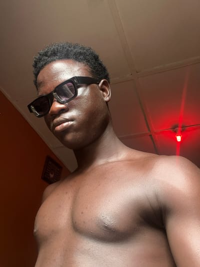 A low-angle, close-up shot of a shirtless, dark-skinned man wearing sunglasses indoors in Mosafejaw, Nigeria. The man is angled towards the viewer and the location contains a red light fixture on the ceiling. A small, dark object rests on an orange surface near the subject.