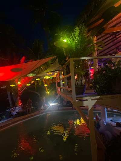At night in Ban Haad Rin, Thailand, a venue is decorated with colorful lights and canvas sails. The lights reflect off a body of water, possibly a pool or pond. Plants and structures are illuminated, with some people visible in the background. The environment suggests an outdoor gathering or party atmosphere.