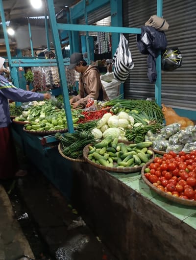 A street-side vegetable stall in Kutawaringin, Indonesia, with a vendor arranging produce on a table. The vendor is a young man wearing a brown hoodie and a black baseball cap. He is arranging green vegetables in a woven basket. Behind the vendor are various other types of vegetables, including cucumbers, green beans, scallions, red chili peppers, green peppers, tomatoes, and gourds. The vegetables are arranged in multiple woven baskets on the table. The vendor is positioned near a building with a blue facade and a metal, slatted roller door. There is a person walking away from the stall, their back is facing the camera. They are wearing a red and blue shirt.