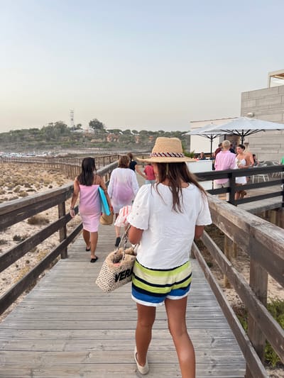 The image displays a coastal scene in a beach area of Almancil, Portugal, during late afternoon or early evening. A group of individuals is walking on a wooden boardwalk that extends from the foreground into the mid-ground. The boardwalk is constructed of weathered wooden planks and features wooden railings on both sides.

In the immediate foreground, a female subject is viewed from behind. She wears a straw hat with a red, white, and blue patterned band, a white short-sleeved top with eyelet embroidery, and horizontally striped shorts in yellow, lime green, blue, white, and black. She carries a light-colored woven tote bag with the word "VERA" embroidered in black. She wears light-colored closed-toe flat shoes.

Ahead of the foreground subject, on the same boardwalk, another female is visible from behind, wearing a pink dress or cover-up and black flip-flops. She carries a light blue object, consistent with a small boogie board or a tightly rolled towel, under her left arm and a straw bag in her right hand. Behind her, a third female, also viewed from behind, wears a translucent white or light pink long cover-up. Further along the boardwalk, additional individuals are observed, all moving in the same direction.

To the right of the boardwalk, a modern, angular building with a light grey, possibly concrete or stone, facade is present. This structure functions as a restaurant or beach club and includes an outdoor patio area. The patio is furnished with multiple white outdoor umbrellas and tables. Several individuals are visible on the patio, standing and socializing. One person wears a pink long-sleeved shirt and light-colored shorts. Another female, in a white dress, holds a glass.

The environment surrounding the boardwalk consists of sandy terrain with sparse, low-lying, dry vegetation, characteristic of coastal dunes. In the mid-ground, beyond the dunes to the left of the boardwalk, a parking area with numerous vehicles is visible. In the far distance, positioned on a vegetated hill, a tall, slender metal lattice structure or tower (potentially a communication tower or part of a lighthouse complex) stands adjacent to a white building. The sky is light and hazy, suggesting twilight conditions.