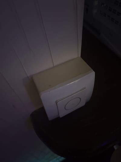 A white doorbell is mounted on a wooden wall. The doorbell is rectangular and has a circular button in the center. The wall is painted white and has vertical lines. The doorbell is located in a dimly lit room. The background is dark and out of focus. The image appears to be taken from a low angle.