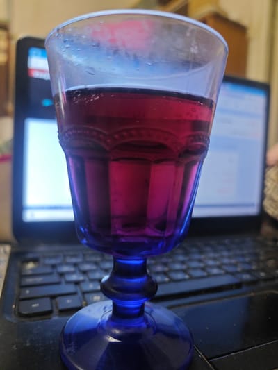 A close-up, low-angle shot shows a clear glass goblet filled with a deep red liquid. The goblet has a blue, decorative base and stem. Condensation is visible on the outside of the glass. The goblet is placed on a laptop keyboard, with the laptop screen blurred in the background. The overall impression is of a casual, indoor setting, possibly a workspace. There is no information in the image to suggest a location in the city of Pushkin, Russia.