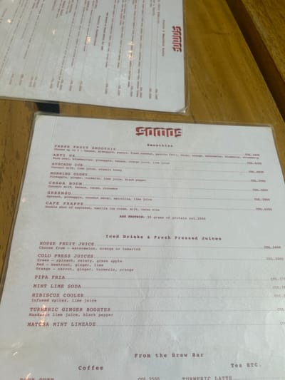 The image shows a close-up of a menu for a cafe called "Smoothie," located in Santa Fe, Costa Rica, at the coordinates 9.644, -85.167. The menu lists various smoothies, juices, and other drinks, including "Fresh Fruit Smoothie," "Iced Drinks & Fresh Pressed Juices," "House Fruit Juice," "Cold Press Juices," "Pipa Fria," "Mint Lime Soda," "Hibiscus Cooler," "Infused Spices, Lime Juice," "Turmeric Ginger Booster," "Mandarin Lime Juice, Black Pepper," and "Matcha Mint Limeade." The prices for each drink are listed in colones, the currency of Costa Rica. The menu is printed on a white paper with red font and is sitting on top of a wooden table surface.