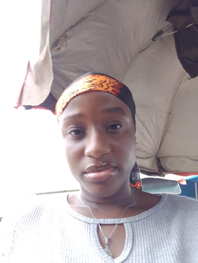 A medium-shot photograph shows a woman with dark skin, wearing an orange patterned headscarf, a gray ribbed shirt with a small cutout at the neck, and a silver necklace. She is positioned beneath a weathered gray market umbrella, suggesting an outdoor location. The background is softly blurred. The setting is suggested to be in the city of Umuahia, Nigeria.