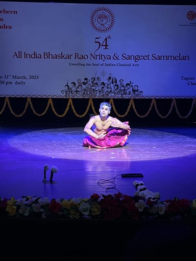 The image shows a stage with a single male dancer performing in front of a backdrop. The backdrop is a white banner with text that reads "54th All India Bhaskar Rao Nritya & Sangeet Sammelan Unveiling the Soul of Indian Classical Arts", as well as a smaller graphic of a circle containing multiple faces.  The stage is dark blue and has an ornate pattern of gold chains hanging down at the front edge. The stage is lit by a single bright spotlight. The dancer is sitting on his knees, wearing a pink garment and a necklace. He is looking directly at the viewer.  The dancer is surrounded by a few loose flowers. The stage is located in the Tagore Theatre at the coordinates 30.733, 76.789 in Chandigarh, India. There are two microphones with white covers on the stage. The scene is dimly lit, suggesting a stage setting. The image was likely captured during a performance.
