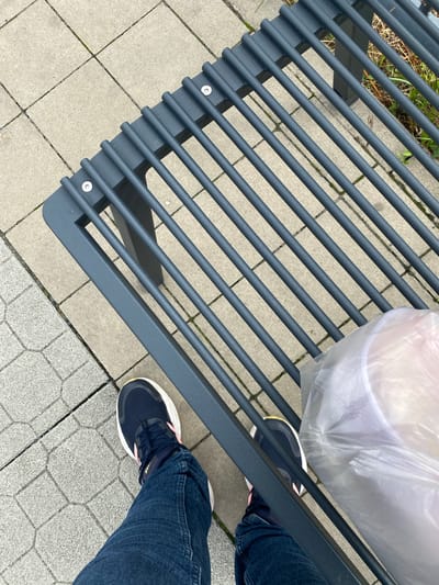 A close-up, overhead shot shows a person's legs and feet standing next to a dark grey slatted park bench on a paved area in Lahošť, Czechia. The person is wearing dark blue jeans and dark blue athletic shoes with pink accents. To the right of the person's feet, a clear plastic bag containing white objects sits on the bench. The ground is composed of rectangular grey paving stones with dark grout lines. Some sparse green vegetation is visible to the right of the bench.
