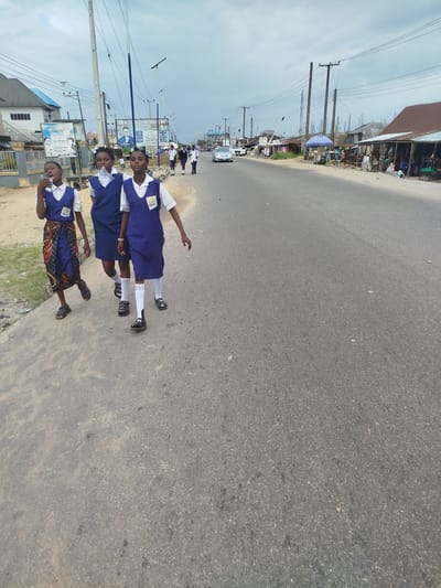 Three young female students in school uniform, consisting of white shirts and blue jumpers, walk along the side of a paved road in Yenegoa, Nigeria. The student on the left is wearing a colorful wrap skirt over her uniform. They appear to be walking away from the camera. The road is bordered by a sandy shoulder and a line of buildings on the left side, including a white building with a blue roof. On the right side of the road, there are various structures, including shops with corrugated metal roofs, some of which appear to be market stalls. Several cars are visible on the road, and utility poles with numerous wires are present along the roadside. The sky is overcast with clouds.