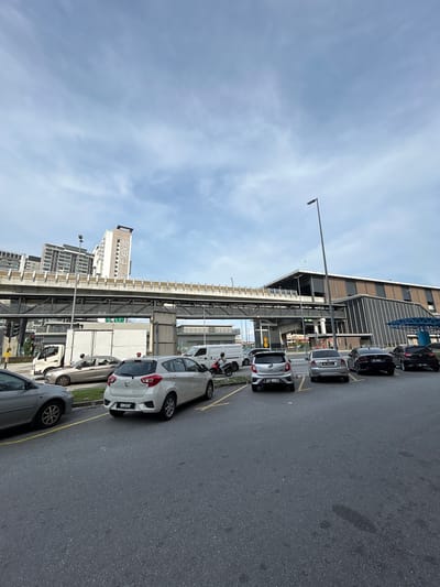 The image shows a street scene in Subang Jaya City Council, Malaysia, with several cars parked and moving. In the foreground, a grey car is partially visible on the left, followed by a white hatchback with license plate VBN 5087. To its right are two sedans, a silver one with license plate WC 6882 Q and a black one with license plate WTV 8534, and another black car is partially visible on the far right. In the background, a large, modern building complex is visible, with an elevated walkway or bridge connecting different sections. A truck is stopped on the left, with a traffic light overhead. A white van is also visible, and a person is riding a motorcycle on the right side of the image. The sky is partly cloudy, with a street lamp visible in the upper right portion of the frame.