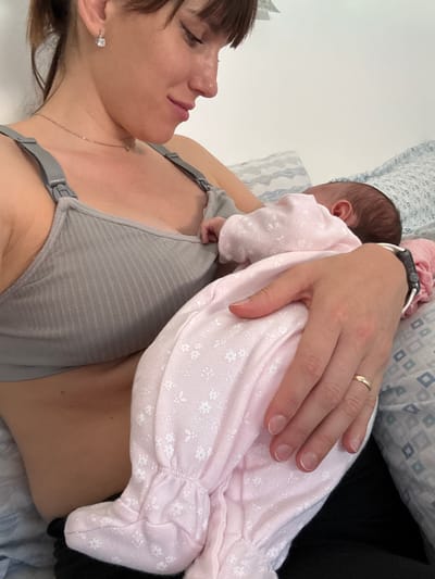 A woman is lying down while holding a baby on her chest. The woman is wearing a grey nursing bra, a delicate gold necklace, and earrings with cubic zirconia. The baby is wearing a pink, floral-patterned onesie. The woman is wearing an Apple Watch on her right wrist and a gold wedding band on her ring finger. They are in a domestic setting, on a bed with pillows. It's impossible to confirm the location in the city of Aprilia, Italy, from this image alone.