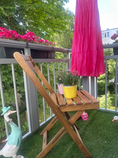 The image shows a balcony with a wooden chair, a pink dress hanging, and potted plants. A bright pink dress is draped over a railing. Next to it, a small yellow pot holds flowering plants, with a pink pot underneath. A wooden chair is positioned on the balcony. On the grass, a pair of pink sandals are visible, along with decorative garden ornaments, including a goose in a hat and jacket and another goose ornament. The setting appears to be a sunny day with lush greenery visible in the background, suggesting a residential balcony. The image is oriented vertically, with the balcony railing running diagonally across the frame.