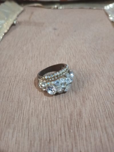 A single ring is depicted resting on a light-brown wooden surface in Chandragiri, Nepal. The ring features a dark-toned metal band adorned with multiple clear, colorless faceted stones arranged in a prominent central cluster and along two parallel rows on the upper part of the band. The wood grain on the surface beneath the ring runs predominantly vertically. The background elements are out of focus, showing blurred light and dark areas. No individuals are present. No actions or interactions are occurring.