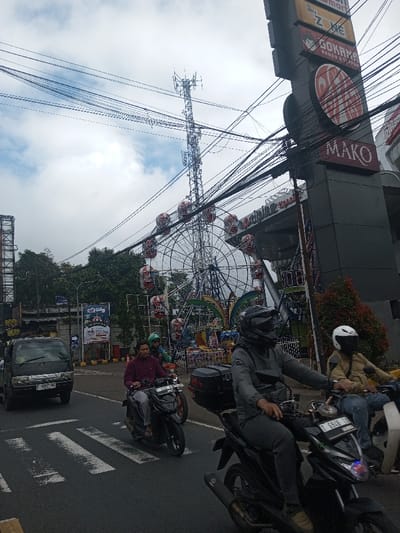 An urban street scene captures a low-angle shot of a Ferris wheel with red and white cars behind a tangle of power lines. Three motorcyclists ride on the street. A black truck sits at a crosswalk, while a multi-tiered building stands tall to the right with several store signs, one reading "MAKO". A cell tower rises in the background under an overcast sky.