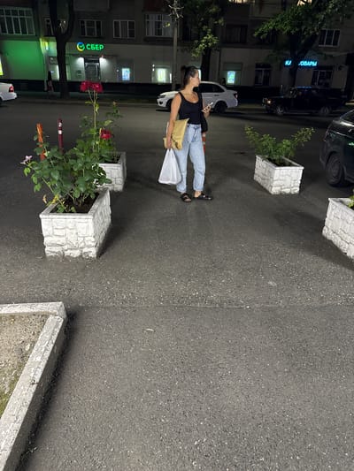 This media file captures an outdoor scene at night, likely in Vladikavkaz, Russia, given the location context. The primary subject is a young woman standing on an asphalt surface, holding a shopping bag and a phone. She is dressed in a black tank top and light blue jeans, wearing black sandals. Her posture and the phone in her hand suggest she might be waiting or simply browsing on her device.

The setting appears to be a street or walkway in front of a building. Several large, white, rectangular planters filled with greenery are visible, some with flowering plants. One planter in the foreground contains red roses and smaller pink blossoms, indicating a mild climate or a well-maintained area. Behind the woman and the planters, a building with multiple windows is dimly lit, hinting at interior lights. A prominent sign with green and yellow lettering, reading "СБЕР" with a circular logo, is clearly visible on the building, identifying it as a Sberbank branch, a major Russian financial institution. Trees with dark foliage are also present, contributing to the nighttime ambiance. The overall impression is one of an urban environment at night, possibly a quiet commercial or residential street. The lighting is artificial, casting soft shadows and illuminating the scene. There is no discernible indication of the weather.