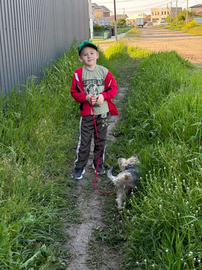 A young boy stands on a dirt path with tall grass on either side, holding a red leash attached to a small dog that stands among the grass. The boy is wearing a green cap, a red jacket, and brown pants. To the left of the boy is a gray metal fence. In the background, there are houses and a road. The location is Rossiyskiy, Russia.