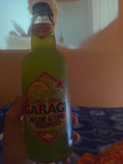 A hand holds a green glass bottle of "Seth & Riley's GARAGE MELON & LIME MADNESS" beer drink. The label is visible, displaying the brand name, flavor, and "4.5%" alcohol content. The background is blurred, suggesting an indoor setting with warm lighting. The location in Mława, Poland cannot be determined from the image.