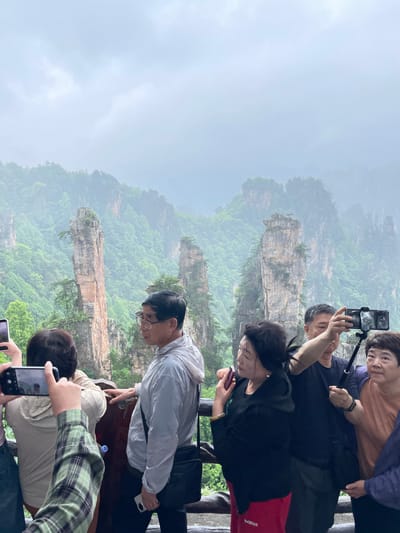 The image shows a group of people at a viewpoint in the city of Wulingyuan, China, likely in the Zhangjiajie National Forest Park. The park is known for its unique sandstone pillars. A layer of fog hangs in the air.
The people are standing near a dark wooden railing. They appear to be tourists, with several holding smartphones and taking pictures of the landscape. Some are looking at their phone screens.
The landscape features numerous tall, slender rock formations covered in greenery. The sky is overcast, suggesting a cloudy day. The overall scene depicts a tourist setting in a scenic natural area.