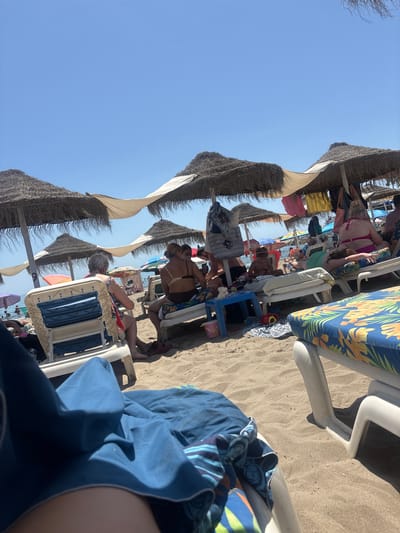 This image captures a sunny day at a beach in Arroyo de la Miel-Benalmádena Costa, Spain. Several thatched umbrellas provide shade over rows of empty and occupied beach chairs. People are seen relaxing on lounge chairs, some reading, others talking, and some appear to be sunbathing. One person in the foreground is covered by a blue towel on a lounge chair. Children are also visible, with one child sitting on a beach chair and playing with something on a small blue table. Other beachgoers are in the background, some closer to the water's edge. The sandy beach is dotted with beach chairs, towels, and small tables. The sky is clear and blue, indicating fair weather.