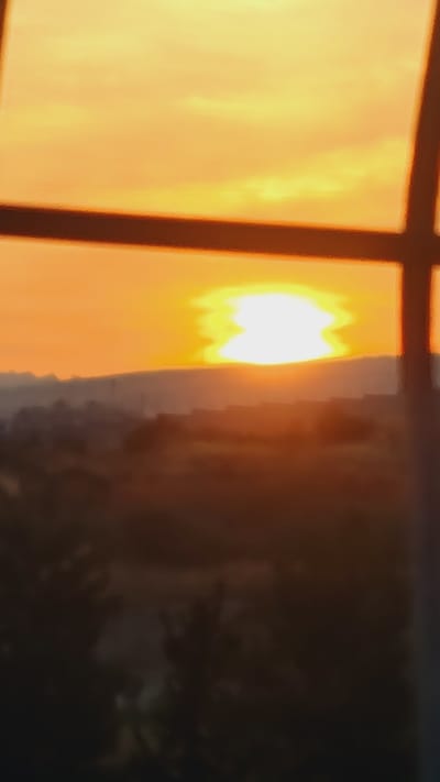 A blurry image shows a bright, orange sunset seen through a window with a dark frame. The area to the right of the sunset is dark and out of focus, possibly showing vegetation or a structure. The location might be Lachish Regional Council, Israel, but this is not visually discernible from the image.