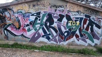 A concrete or stucco wall, situated in Santa Ponça, Spain, is extensively covered in multiple layers of graffiti. The wall exhibits signs of aging and wear, with a prominent brown-tiled or concrete roof edge visible along its top. The lower portion of the wall meets a narrow strip of unkempt green grass, which transitions into a gravel and dirt ground in the foreground.
The graffiti consists of various styles and applications. A large, multi-colored graffiti piece dominates the central and right sections of the wall, featuring complex lettering in shades of dark purple, green, dark blue, and white outlines, partially obscuring earlier tags. A yellow tag spelling "TOY" with a circled 'Y' is present within this central artwork. To the left, a distinct graffiti piece features pink and black outlines with red lettering, including the word "JESUS." Numerous smaller, overlaid tags and throw-ups are discernible across the entire wall, in colors such as yellow, black, pink, and light blue. Examples of visible tags include "OKAS," "KEO," "MR4," and "4JO5Z." Above the roofline, sparse tree foliage is visible against an overcast sky. No individuals are present in the scene.