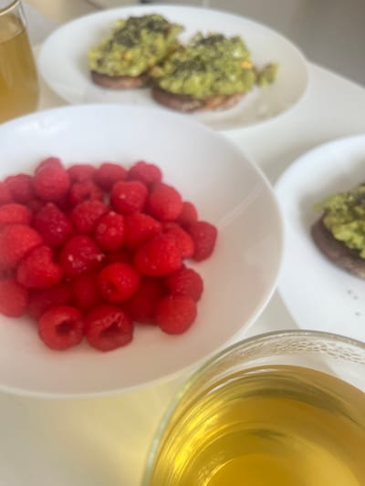 The image presents a close-up, overhead view of a light-colored surface featuring a meal consisting of raspberries in a white bowl, avocado toast on two separate white plates, and a glass of tea. The raspberries are bright red and clustered together in the center of the bowl. The avocado toast appears to be topped with a green spread. The tea in the glass is a light amber color. The scene seems to be a breakfast or light meal setting. While the city is specified, the exact location within Mogán, Spain is not discernible based on the provided image.