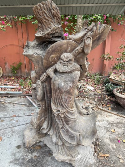 An weathered wooden sculpture, depicting a bald, bearded figure with an angered expression and furrowed brow, stands centrally on concrete ground in Đăk Mar, Vietnam. The figure wears flowing robes and carries a large, circular, segmented object with radial lines on its back, held by its left hand over the left shoulder. The sculpture exhibits a dark, aged wood grain with visible cracks and areas of lighter discoloration.
The immediate ground surface is composed of grey concrete, partially covered with scattered dry leaves and small debris. To the left of the sculpture's base, several thin metal rods or pipes are laid horizontally on the ground.
In the background, a solid reddish-orange wall extends across the frame. Above and along the wall, green foliage with vibrant pink bougainvillea-like blossoms is visible. To the far right, a brown ceramic pot containing a small, pruned tree (possibly a bonsai) is situated on a patch of dirt and sparse greenery. Nearby, a wooden pallet or crate, covered with debris, rests on the ground. An overhead corrugated metal awning or roof is visible at the top of the image, providing partial shade to the scene. No individuals are present in the image.