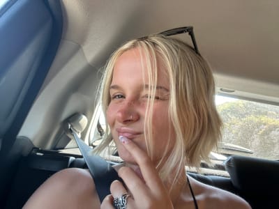 In the city of Elaties Municipal Unit, Greece, a young woman with blonde, shoulder-length hair is seen in the back seat of a car, facing forward and looking at the camera. She is winking, with her right eye closed and her left eye open. Her left hand is raised, with her index finger touching her lips. She is wearing a black top and a silver ring on her right hand. The background shows the interior of the car, including a seatbelt, a headrest, and a window with trees and sunlight visible outside. The image appears to be a selfie, taken from a low angle.