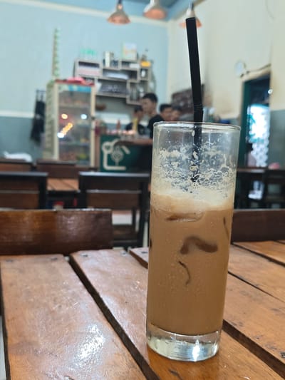 A tall glass of iced coffee with a black straw sits on a wooden table in the foreground. The drink is light brown with foam at the top and several ice cubes visible. The table appears to be made of rough-hewn wooden planks. In the background, several rows of similar wooden tables and chairs are visible, suggesting a cafe or restaurant. Beyond the tables, shelves are stocked with various items, and a few people are blurred in the distance. The lighting is dim, with some lights hanging from the ceiling. The overall scene is casual and appears to be indoors. The location is identified as Blangmetimu, Indonesia.