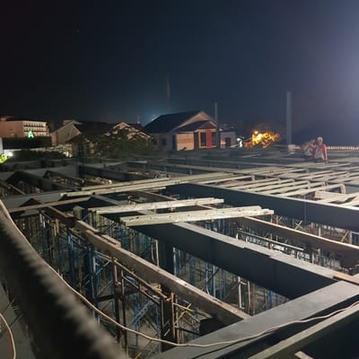 The image depicts a construction site at night in Banda Aceh, Indonesia. Two construction workers, one wearing a red shirt and yellow hard hat, and another in a white shirt and yellow hard hat, are visible near the top of a building structure. They appear to be working at a significant height. The construction site is filled with scaffolding, steel beams, and wooden planks, indicating ongoing work. The surrounding environment is dark, with some artificial lighting from street lamps and the building's interior visible in the background. The time of day is clearly night, and the weather appears to be clear. The perspective of the image is from a low angle, looking upwards towards the workers and the structure. There is no visible text in the image.