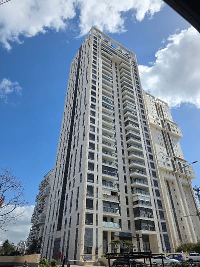 The scene depicts a tall, contemporary residential skyscraper located on Rothschild Street in Rishon LeZion, Israel. The building features a light-toned facade, likely constructed from stone or precast panels, extensively detailed with dark-framed windows and multiple glass-fronted balconies arranged in vertical stacks. Darker vertical elements provide contrast on the building's exterior. The structure ascends into a bright blue sky, partially covered by scattered white cumulus clouds. At ground level, a paved sidewalk fronts the building, where indistinct human figures are visible. Several parked vehicles, including a light-colored sedan and a dark vehicle, are present on the adjacent street. A tall utility pole with overhead lines stands on the far right side of the frame. Portions of an interconnected or adjacent building, exhibiting similar modern architectural styling and balconies, are discernible to the left and rear of the primary tower.