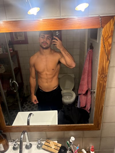 The image depicts a shirtless man taking a selfie in a bathroom mirror. The bathroom has tiled walls, a toilet with the lid open, a sink, and a wooden-framed mirror with a chain for hanging. There is a towel hanging above the toilet and toiletries near the sink. Based on the presence of toiletries, a framed poster, and the presence of clothing, the bathroom likely exists within a private residence. Location in Avellaneda, Argentina, cannot be determined solely from the image.
