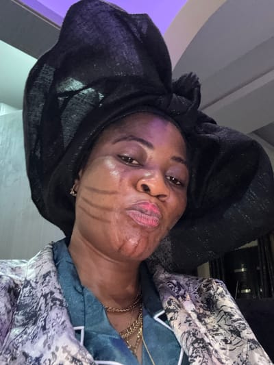 A close-up, selfie-style photograph features a dark-skinned woman from Ajara, Nigeria, wearing a large, elaborate black headwrap and a patterned jacket. The woman has distinct, dark lines drawn on her cheeks, resembling tribal markings, and her lips are pursed. She is wearing multiple gold necklaces. The background is dimly lit and appears to be an indoor setting with a textured wall and a hint of purple lighting from above.
