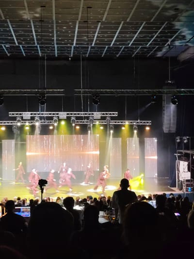 The image depicts a performance on a stage in what appears to be an indoor theater or concert hall. Several performers, dressed in dark outfits, are engaged in dance or choreography on a brightly lit stage. The stage features a large backdrop with a shimmering, abstract design and vertical light columns. The lighting on stage is dramatic, with spotlights and colored washes illuminating the performers. In the foreground, the backs of an audience are visible, seated in darkness, with some individuals holding up phones or cameras. The ceiling of the venue is visible in the upper portion of the image, featuring a grid-like structure with lighting fixtures. The overall scene suggests a live performance event. The location is not specified within the image, and therefore cannot be identified as being in the city of Mykolaiv, Ukraine.